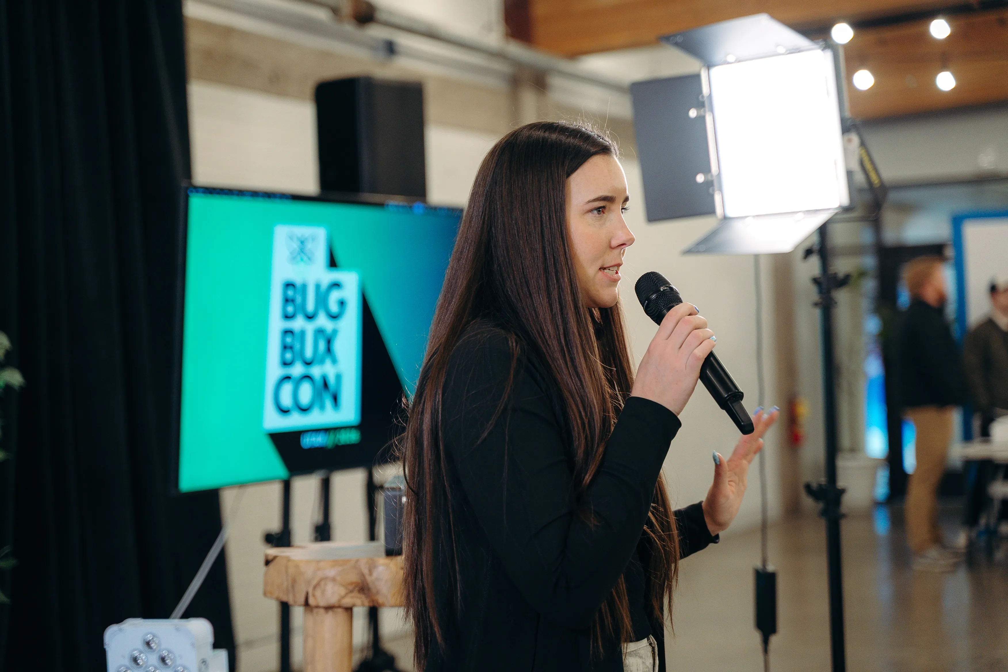 A speaker with a microphone in front of the Bug Bux Con screen